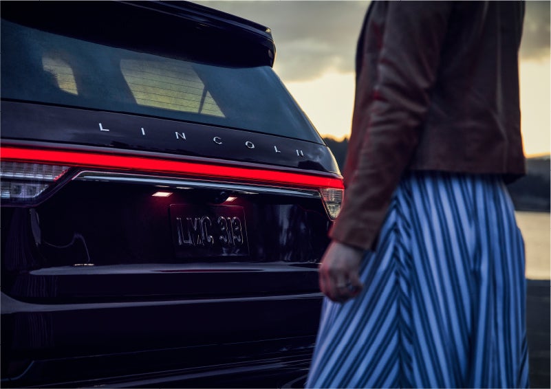 A person is shown near the rear of a 2023 Lincoln Aviator® SUV as the Lincoln Embrace illuminates the rear lights | Irwin Lincoln Laconia in Laconia NH
